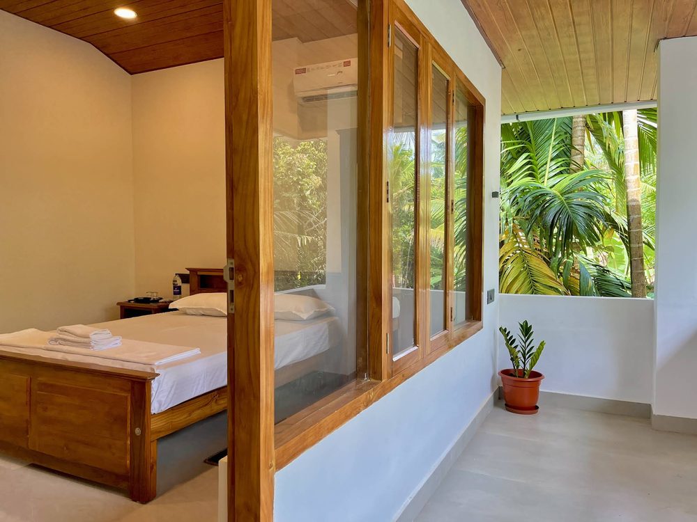 A bedroom at Olam seen through its glass partition, with the verandah and coconut palms beyond