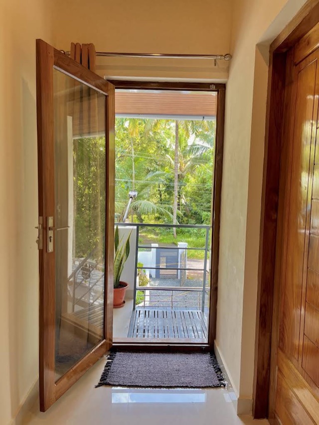 A teak and glass door standing open onto a balcony, doormat on the tiles, palms and a lane visible past the railing