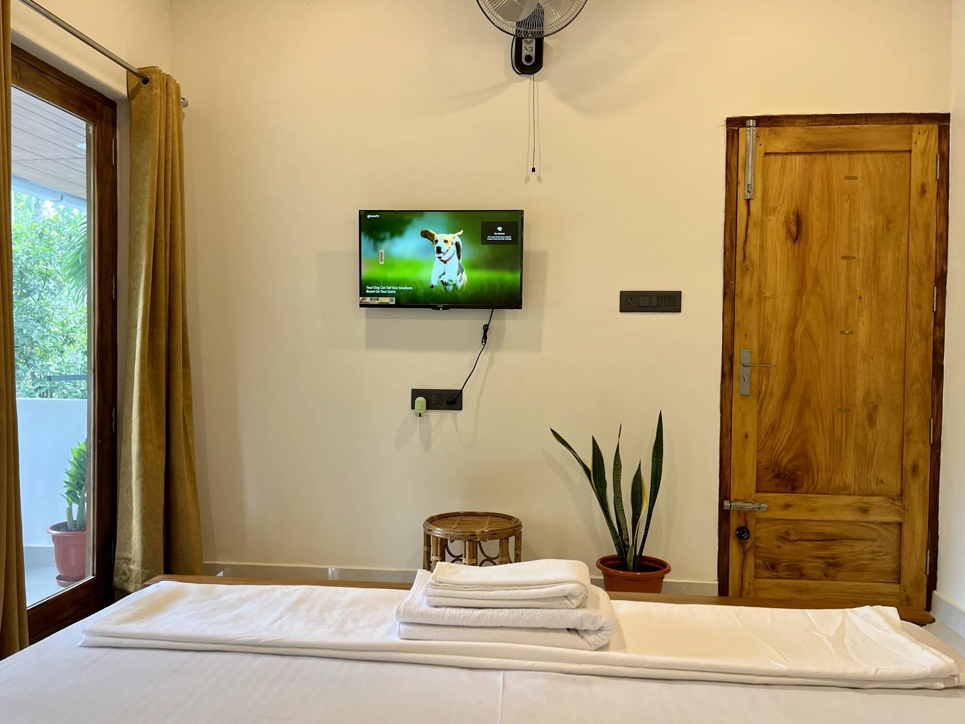 Made bed with folded towels, a wall-mounted television and fan opposite, solid teak door to one side, balcony door to the other