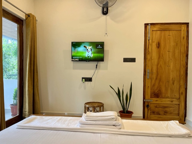 Made bed with folded towels, a wall-mounted television and fan opposite, solid teak door to one side, balcony door to the other